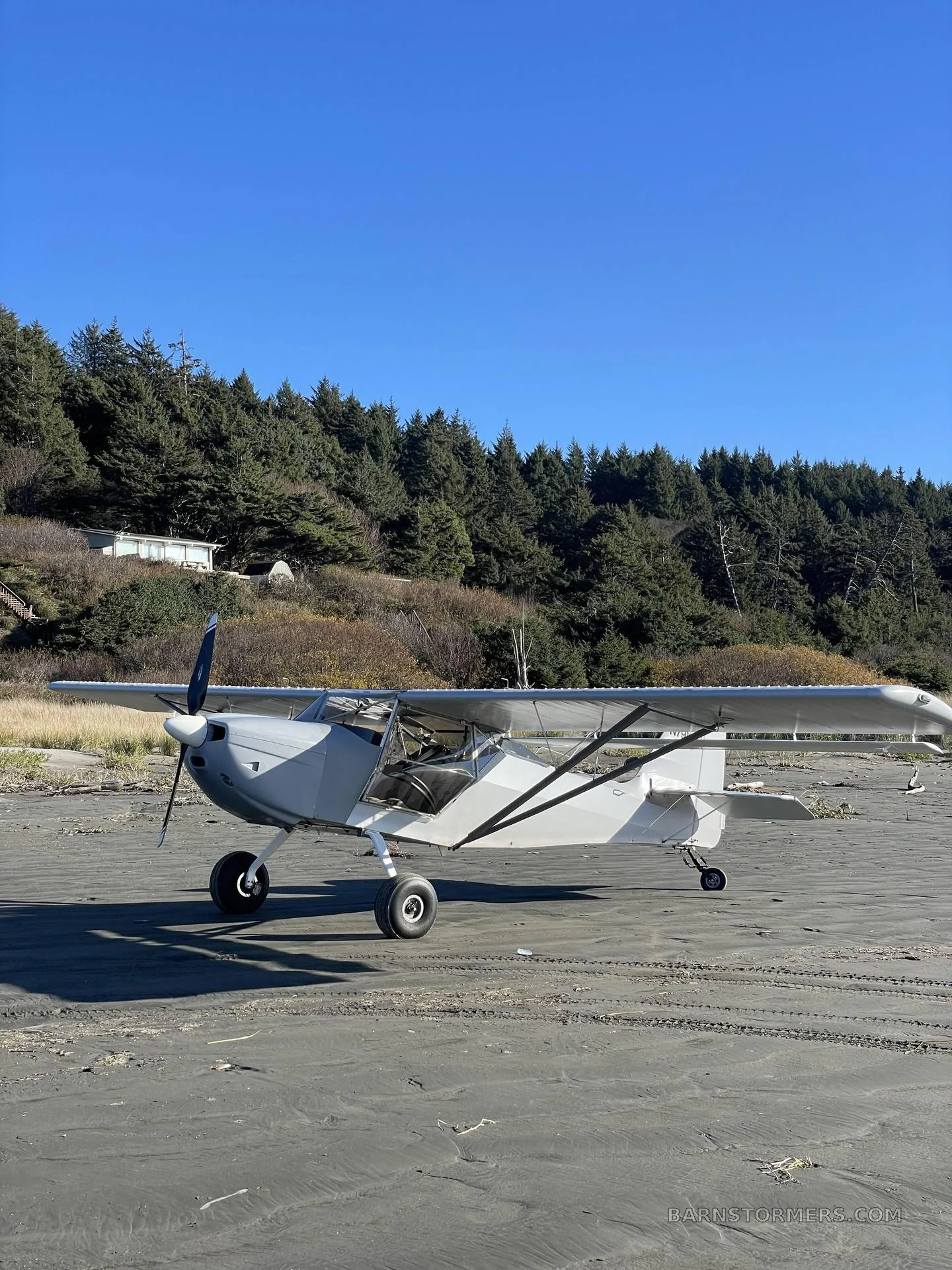 A light grey single-engine aircraft with a high wing and large tires is parked on a sandy beach with a dense green forest on a hill in the background.