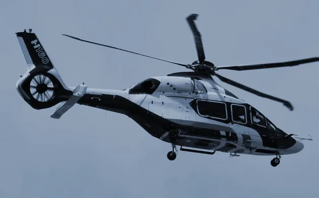 An Airbus H160 helicopter in a black and white livery is captured in flight against a light blue sky.
