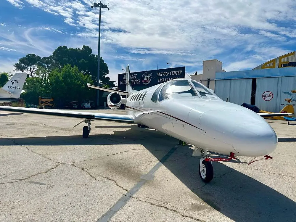 A white Cessna CitationJet aircraft with a red stripe is parked on an airport tarmac under a cloudy sky.