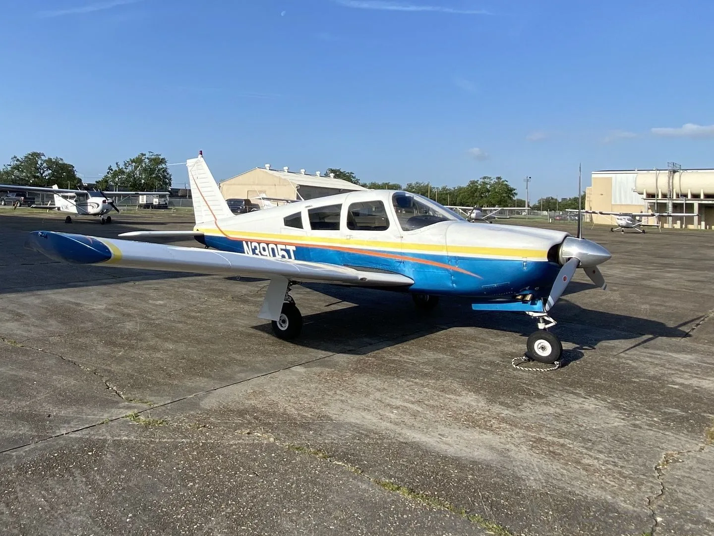 A Piper Cherokee-style aircraft, N3905T, with a white, blue, and yellow paint scheme, parked on a concrete apron on a sunny day.