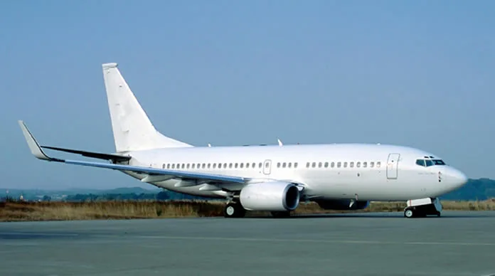 A white Boeing 737 aircraft with a plain livery parked on an airfield under a clear blue sky.