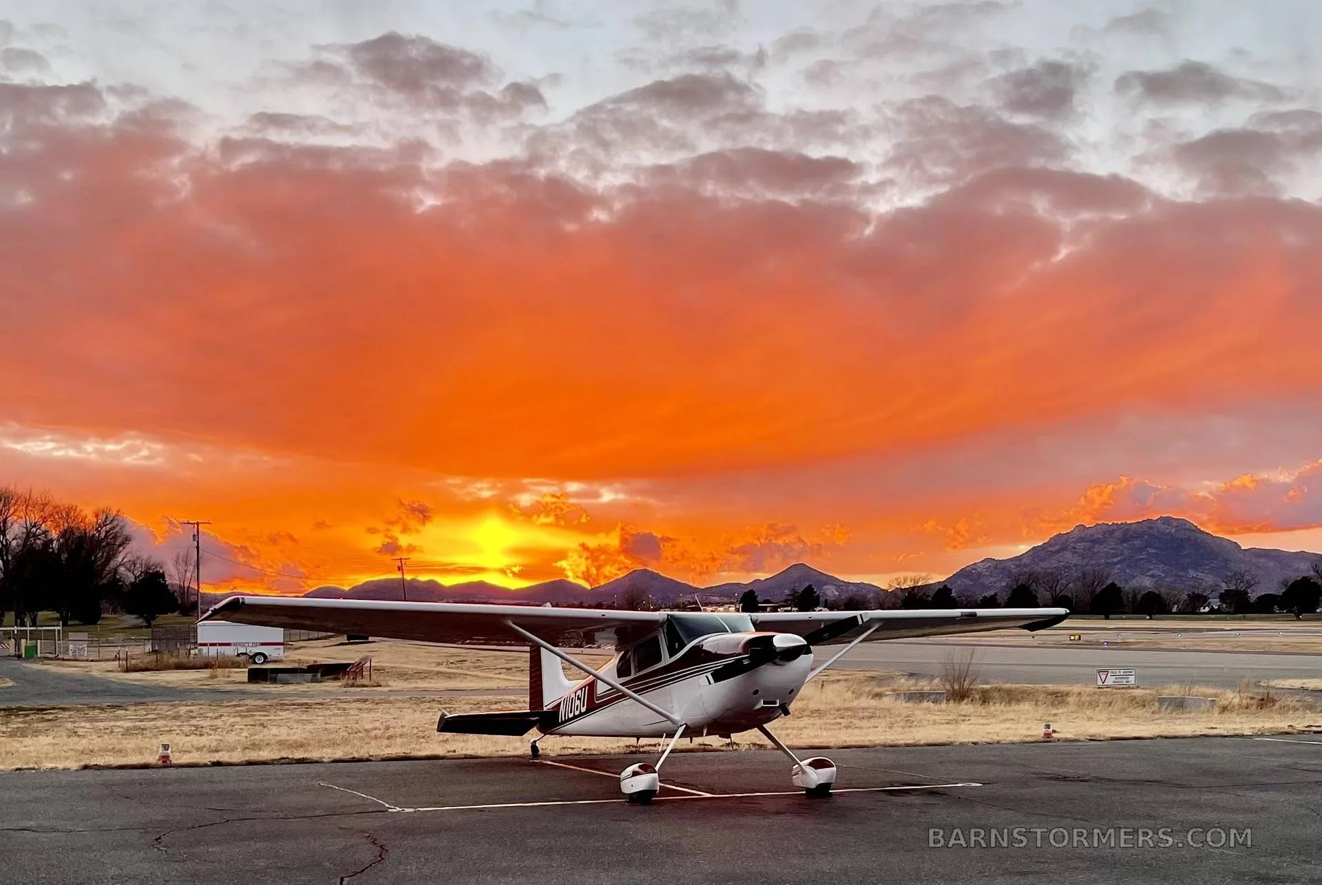 A white and maroon Cessna 182 aircraft (N106U) is parked on an airfield tarmac under a vibrant orange and red sunset sky with mountains in the background.