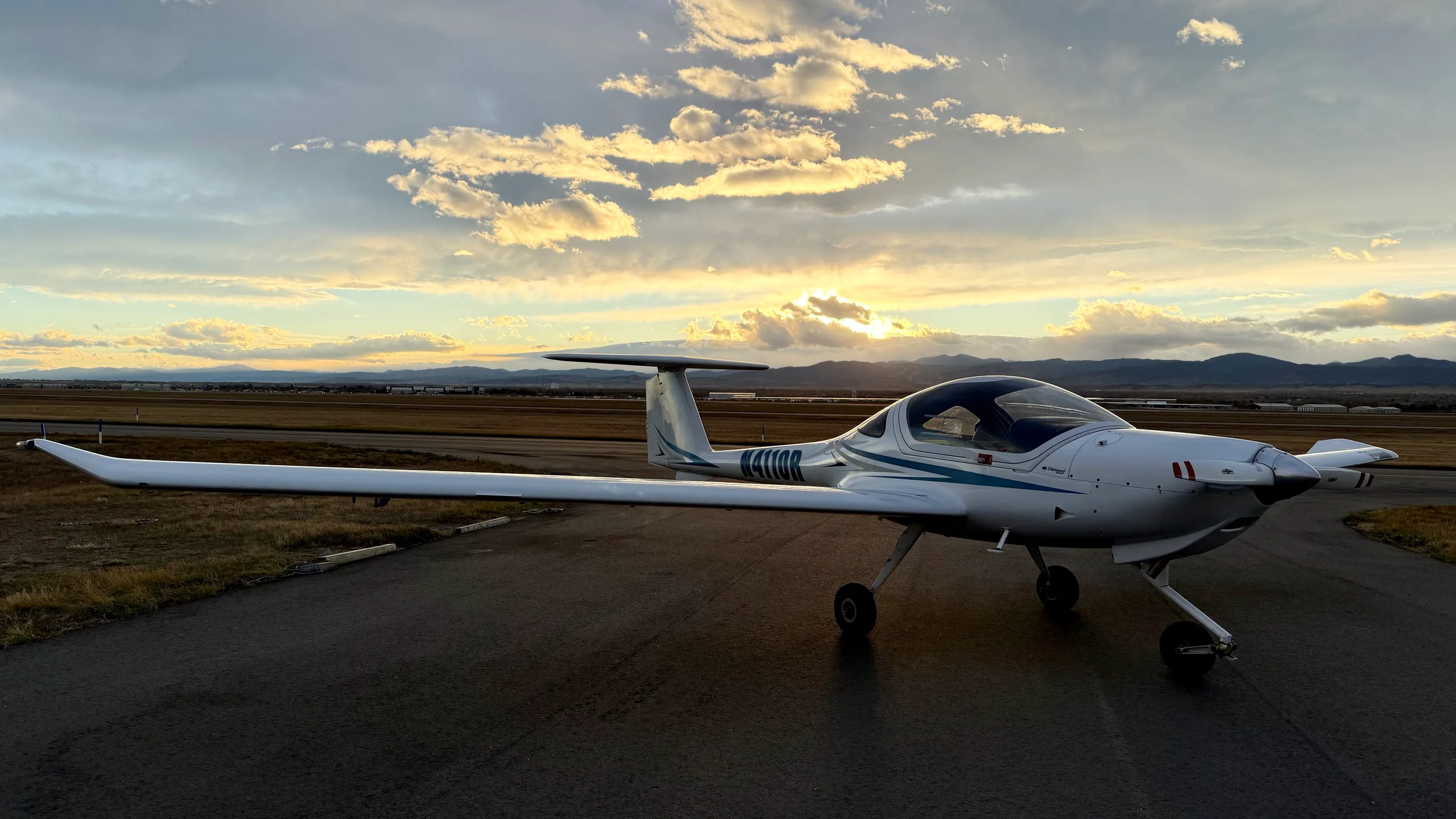 A white Diamond DA20-C1 Eclipse aircraft with blue stripes, registration N4110R, parked on an airfield with the sun setting in the background.