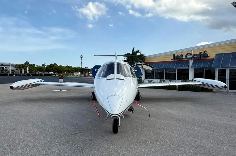 A white Learjet with red and blue stripes, registration N747BW, seen from the front parked in front of a "Jet Café" building.