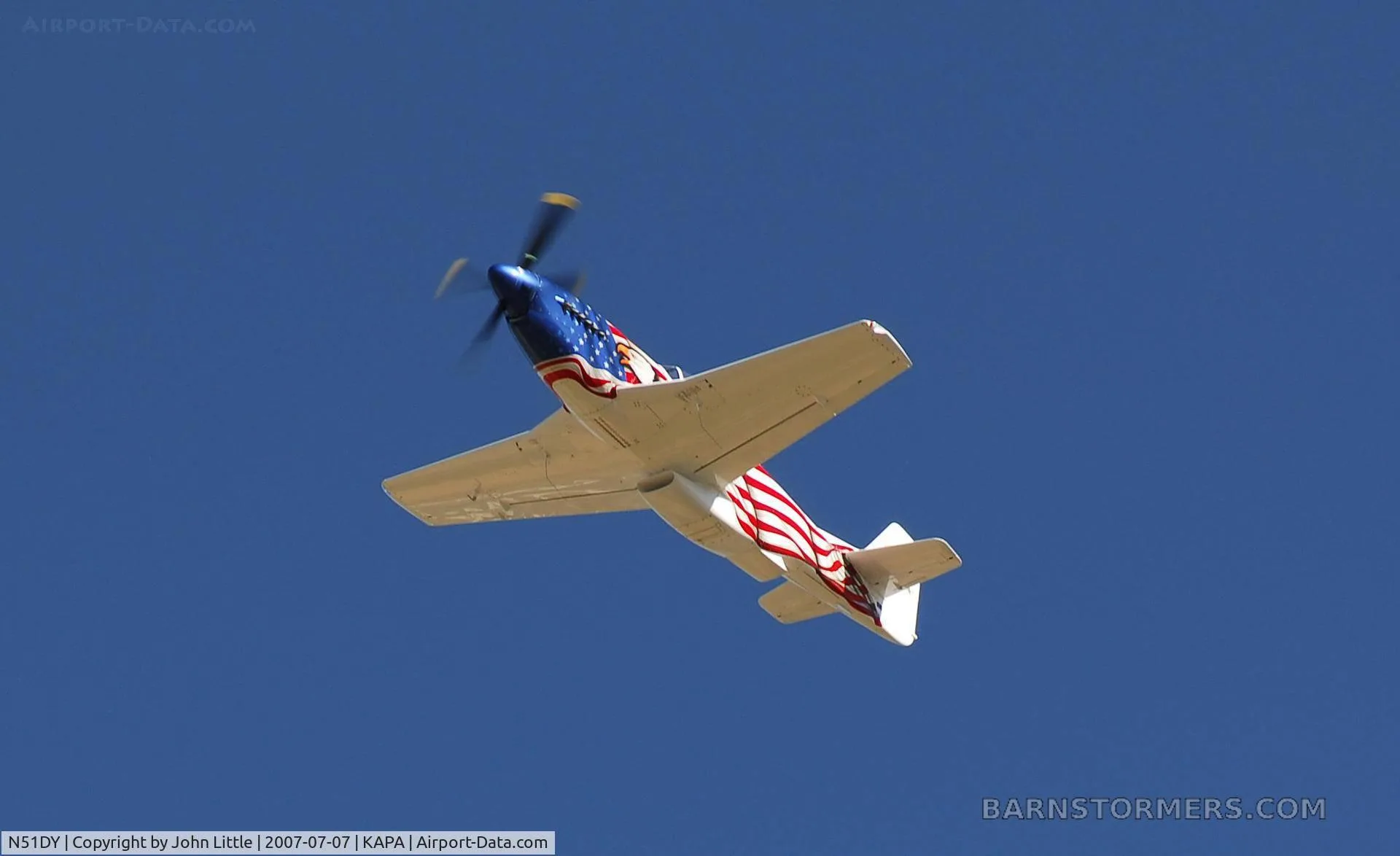 An American flag-painted P-51 Mustang replica flying away, showing its underside against a clear blue sky.