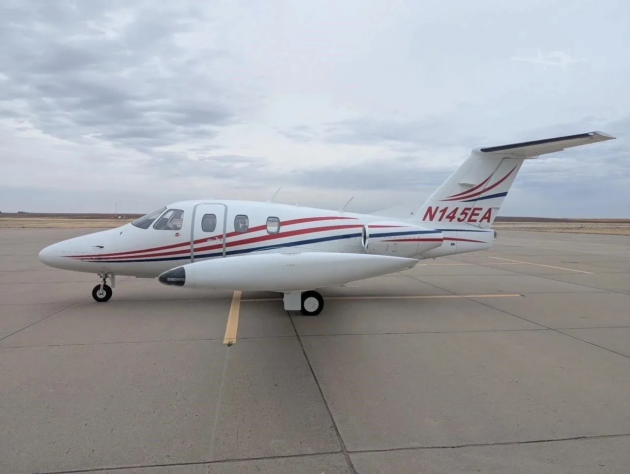 A white Eclipse jet with red and blue stripes, registration N145EA, parked on an airfield from a side view.