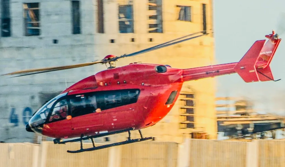 A red Airbus H145 helicopter hovers in front of a large, light-colored building under construction.