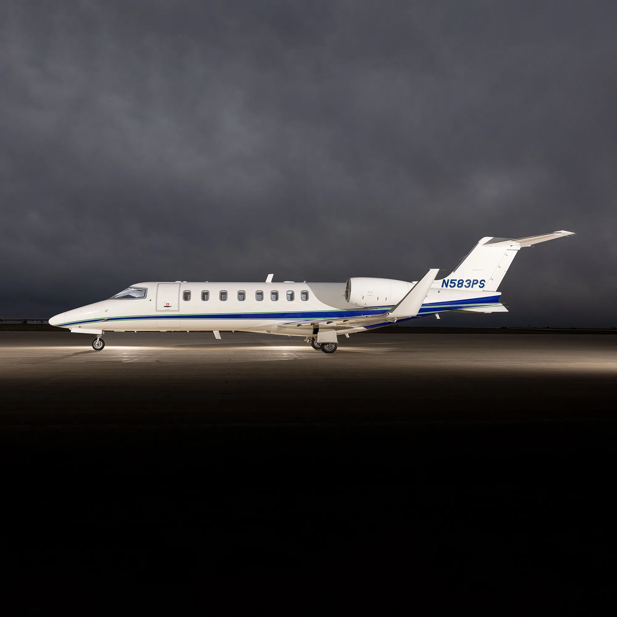 A white Learjet 75 with blue and green stripes, registration N583PS, parked on an tarmac under dramatic cloudy skies.