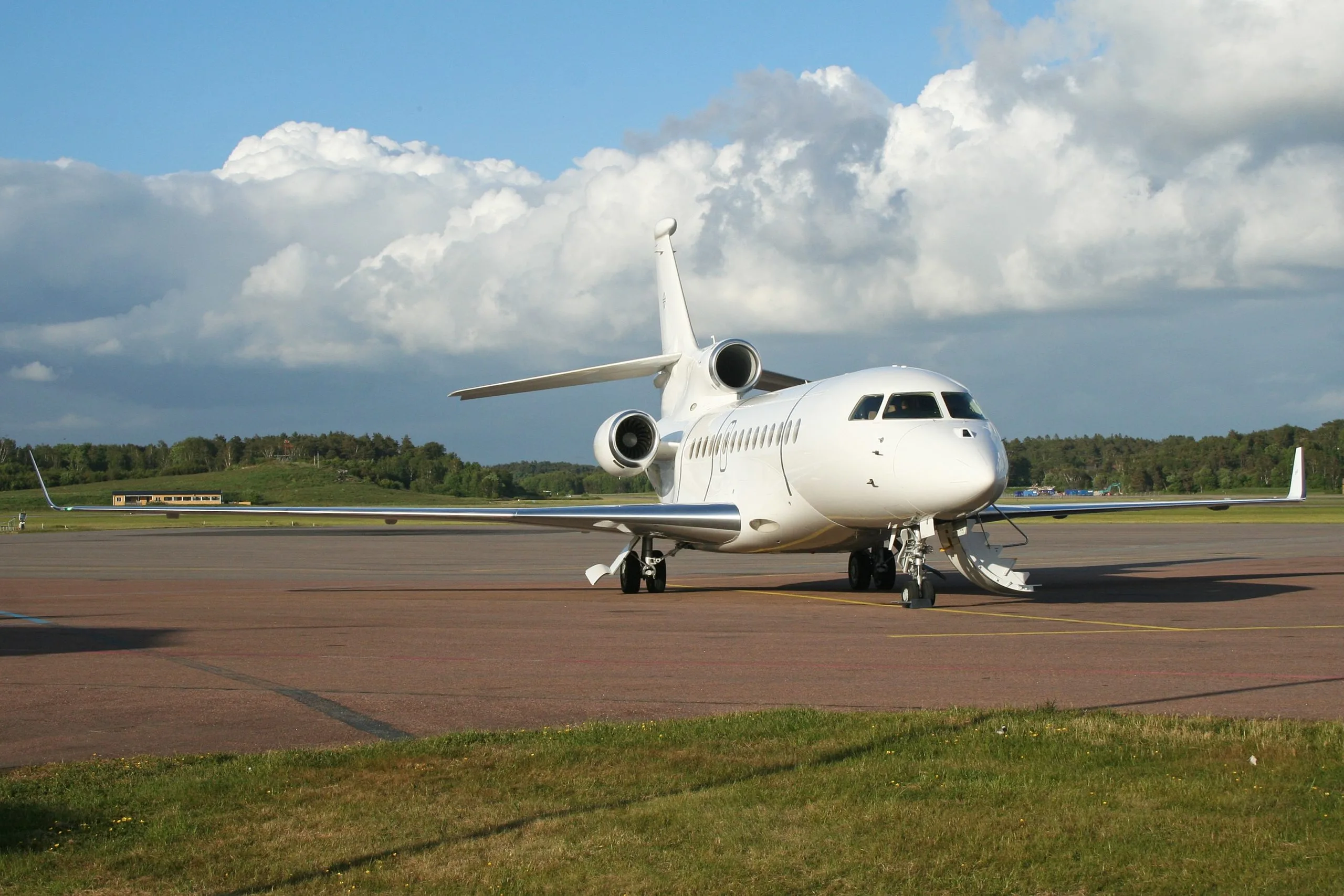 A white business jet with three engines is parked on an airport tarmac under a partly cloudy sky.