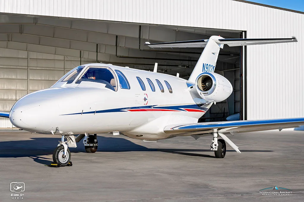 A white Cessna Citation CJ3 (N903GW) with blue and red stripes parked in front of a hangar.