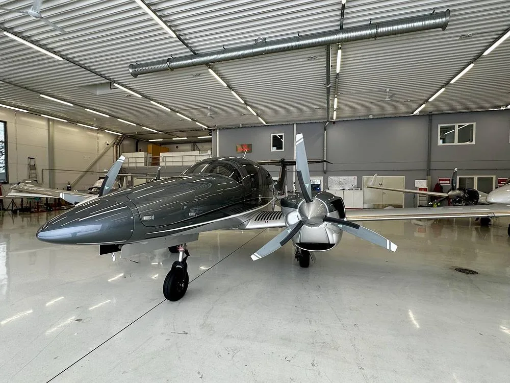 A front-left view of a gray and silver Diamond DA62 aircraft parked in a well-lit hangar.