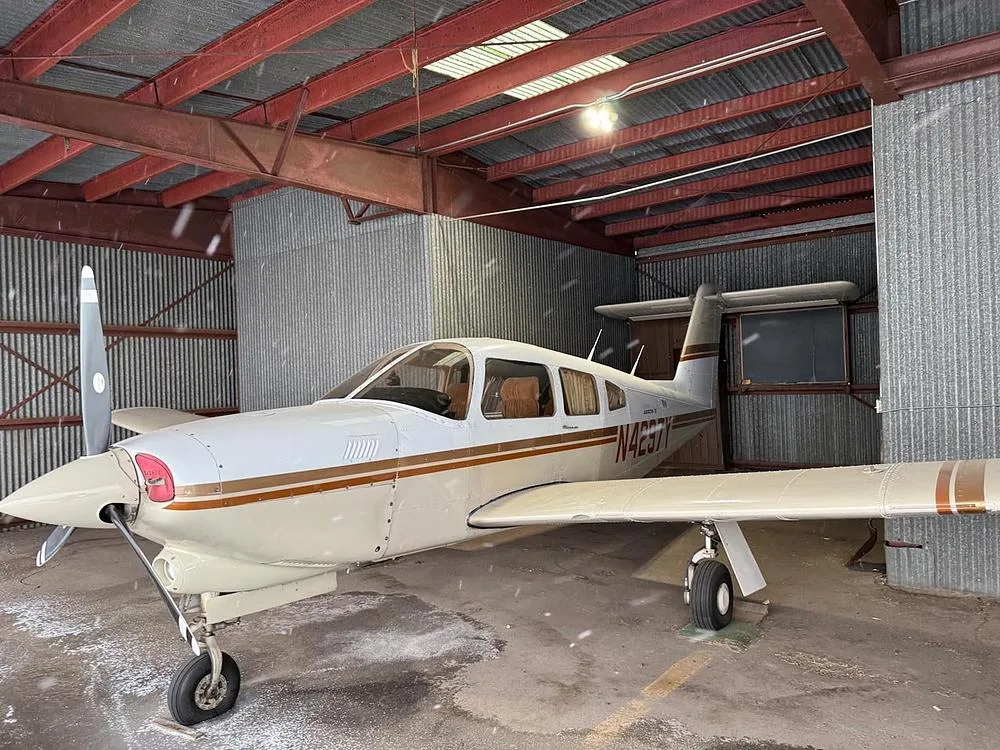 A three-quarter front view of the Piper Arrow IV aircraft, N4297Y, with white and brown livery, parked in a hangar.