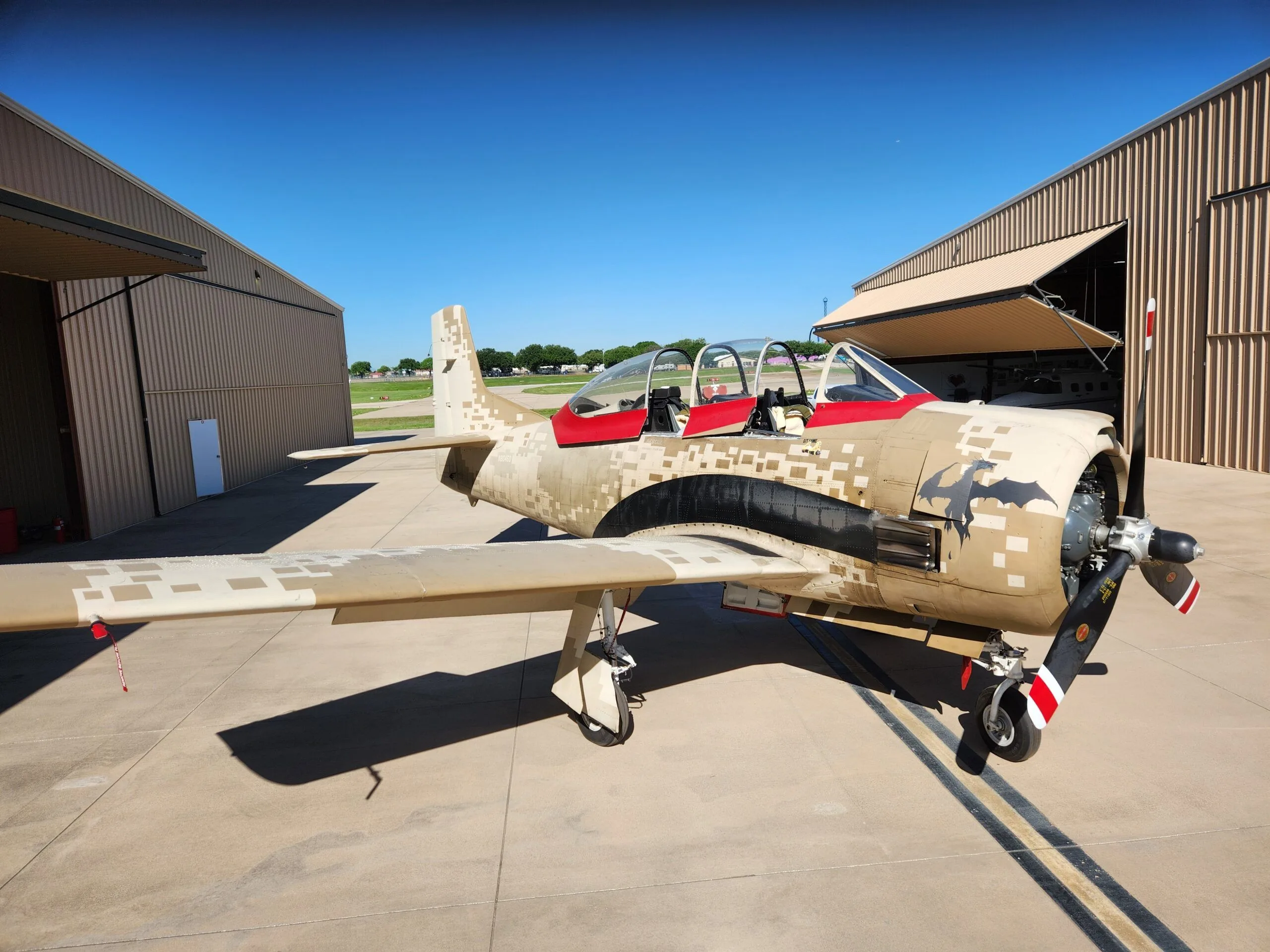 A T-28 Trojan aircraft with a desert digital camouflage paint scheme and red accents is parked on an airfield next to hangars.