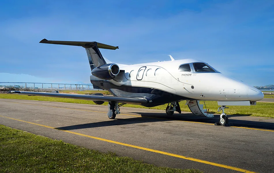A white and dark grey Embraer Phenom 100 business jet is parked on an airfield taxiway under a clear blue sky.