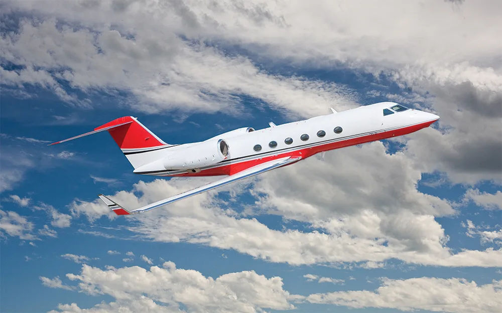 A white private jet with a red stripe on its fuselage and tail flies through a cloudy blue sky.