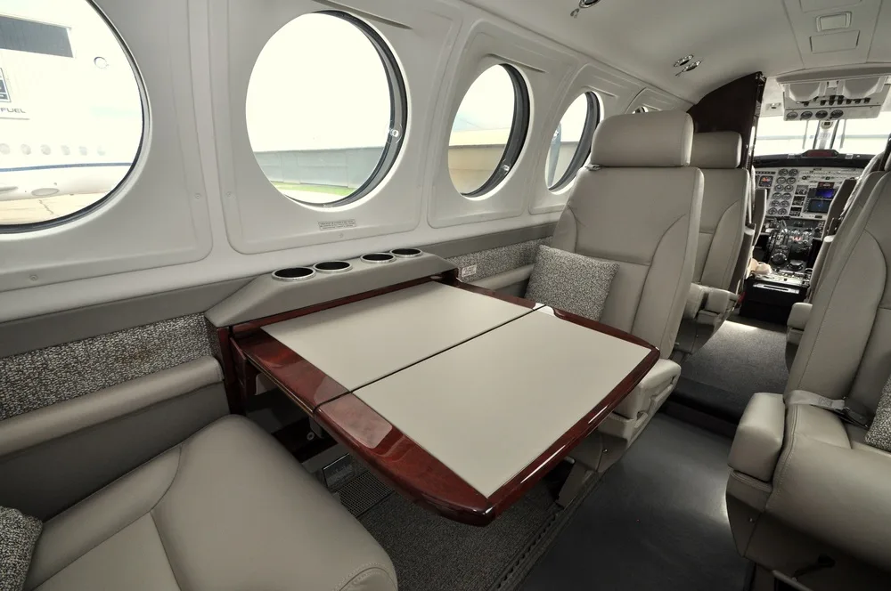 The aircraft's cabin interior featuring a fold-out wooden table and light-colored leather seats, with the cockpit visible in the distance.