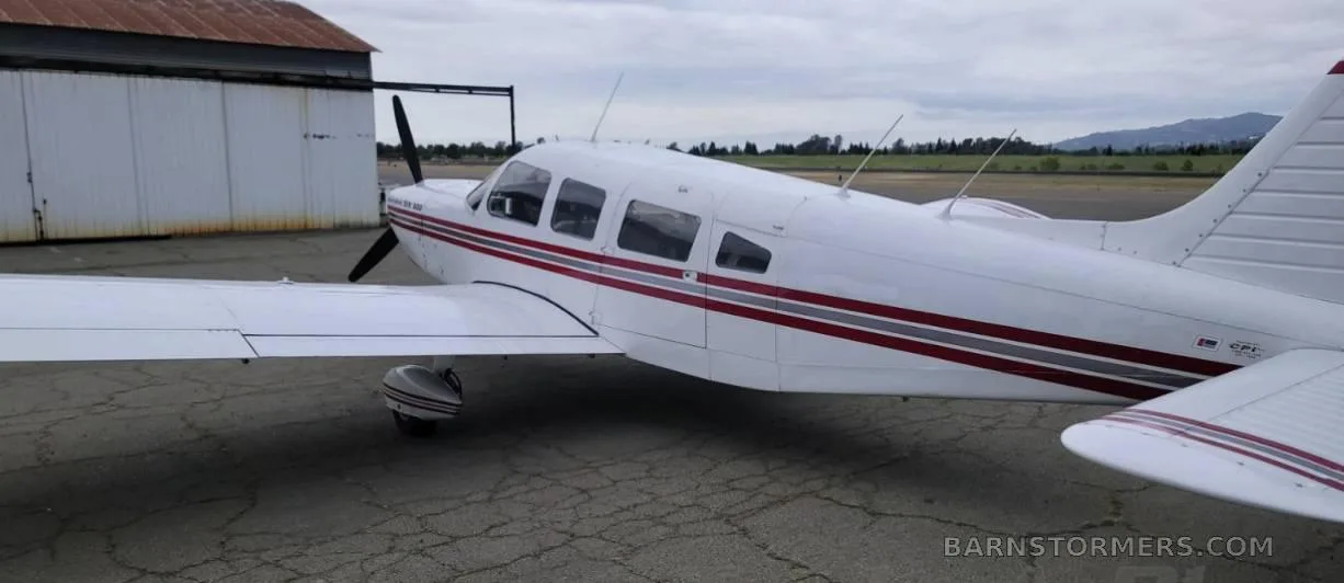 A white Piper Cherokee Six 300 aircraft with red and grey stripes, seen from the mid-left side, parked on an asphalt apron next to a hangar.