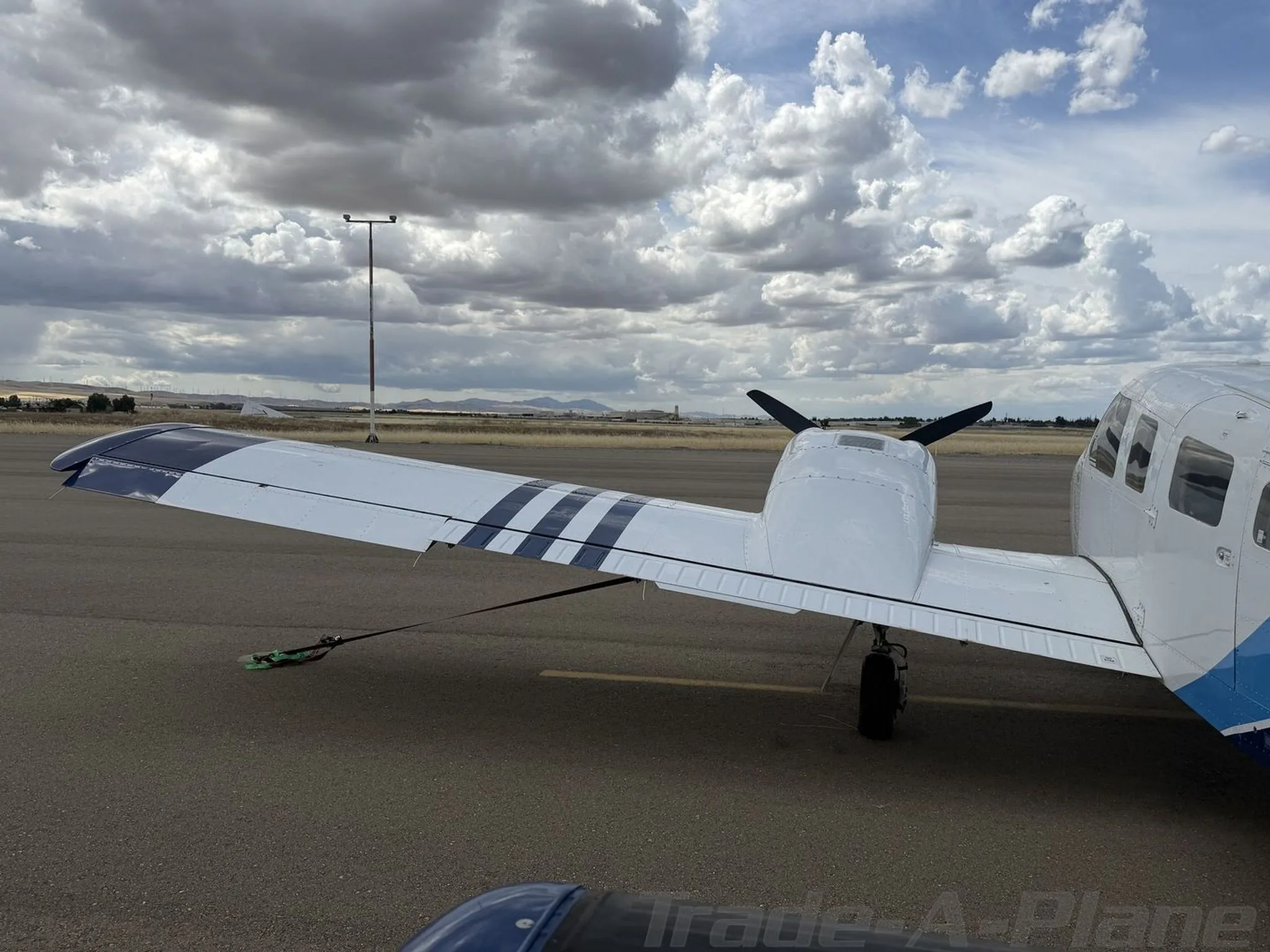A rear-left view of the wing and fuselage of a white Piper Seneca II aircraft with blue stripes on the wing, tied down on an airport tarmac.