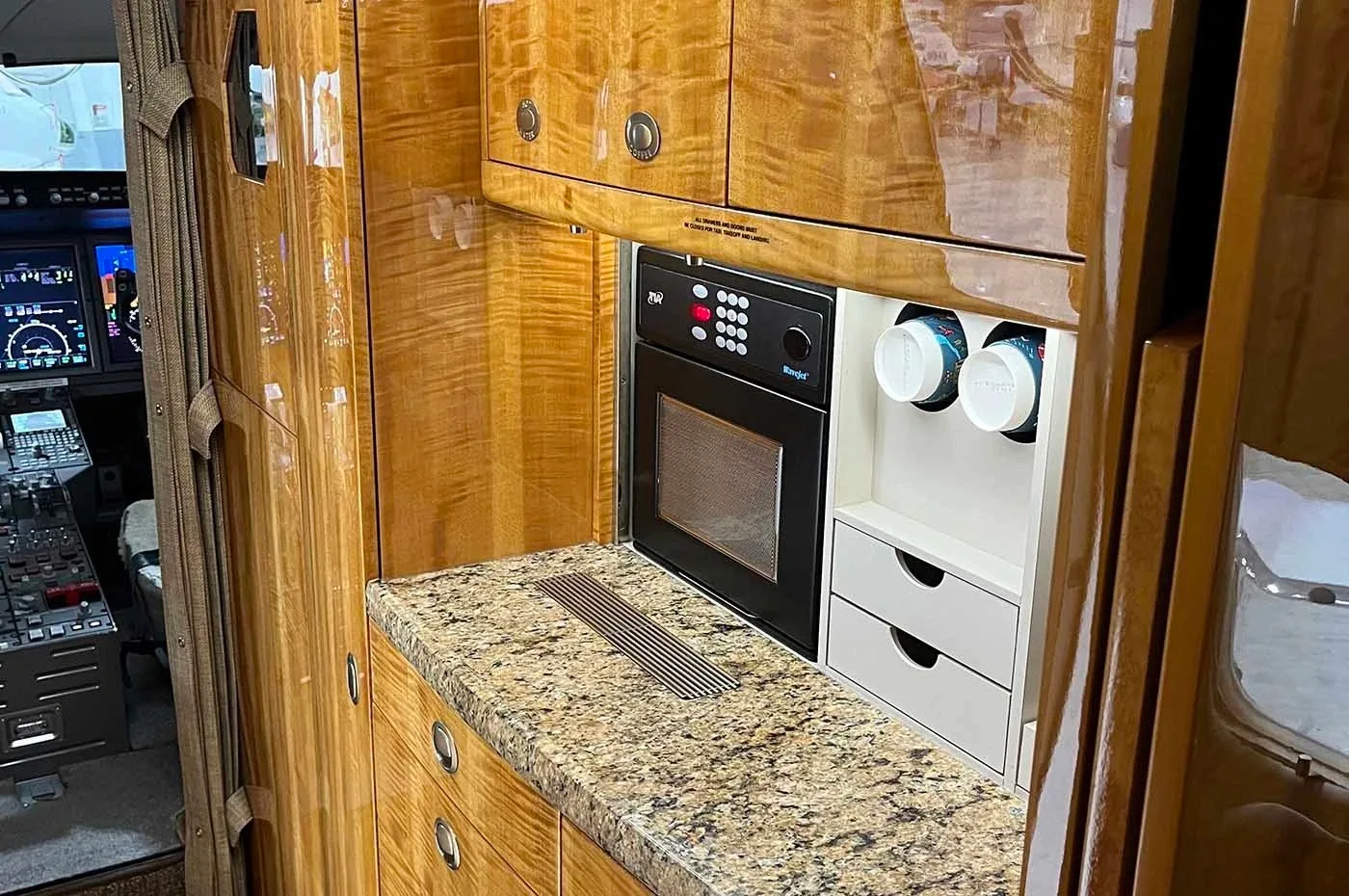 A well-appointed aircraft galley with wood cabinetry, a microwave, and a granite countertop, with the cockpit visible in the background.