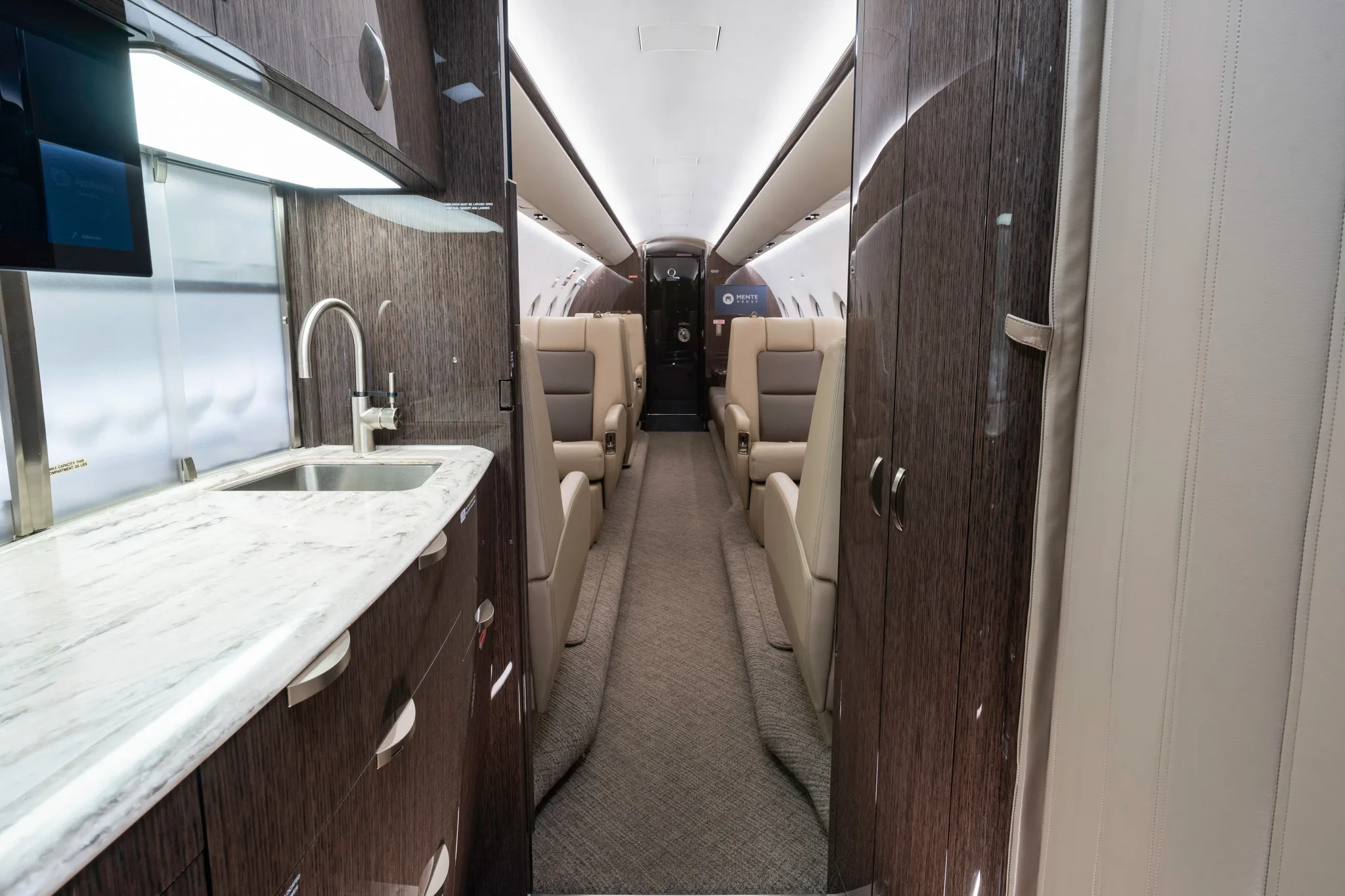 The interior of a private jet, showing a galley area with a sink leading into the main cabin with beige seats.