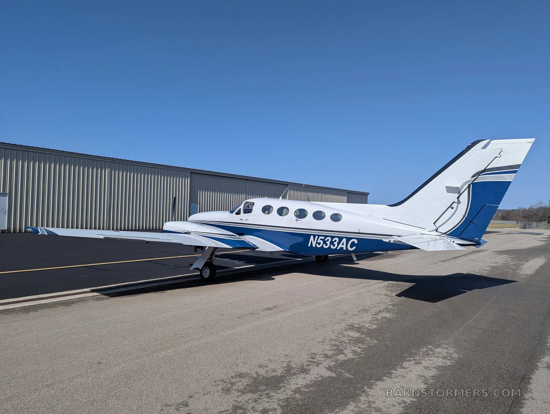 A white and blue twin-engine propeller aircraft (Cessna 414 Chancellor) with tail number N533AC parked on an asphalt ramp next to a hangar.
