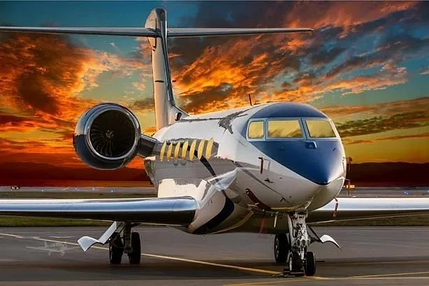 A white and blue business jet with a polished fuselage sits on the tarmac against a vibrant orange and purple sunset.