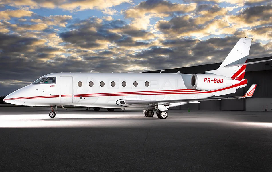 A white business jet with red stripes and registration PR-BBD is parked on a tarmac under a dramatic cloudy sky.