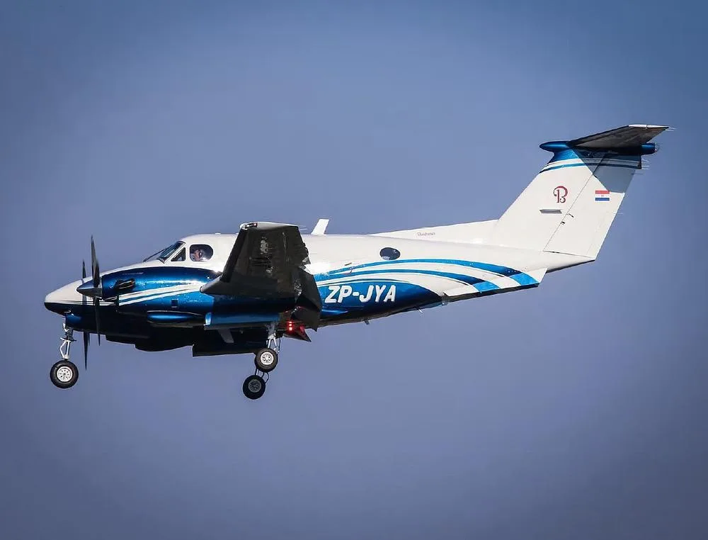 A Beechcraft King Air with blue and white livery, registration ZP-JYA, flying with its landing gear extended against a blue sky.