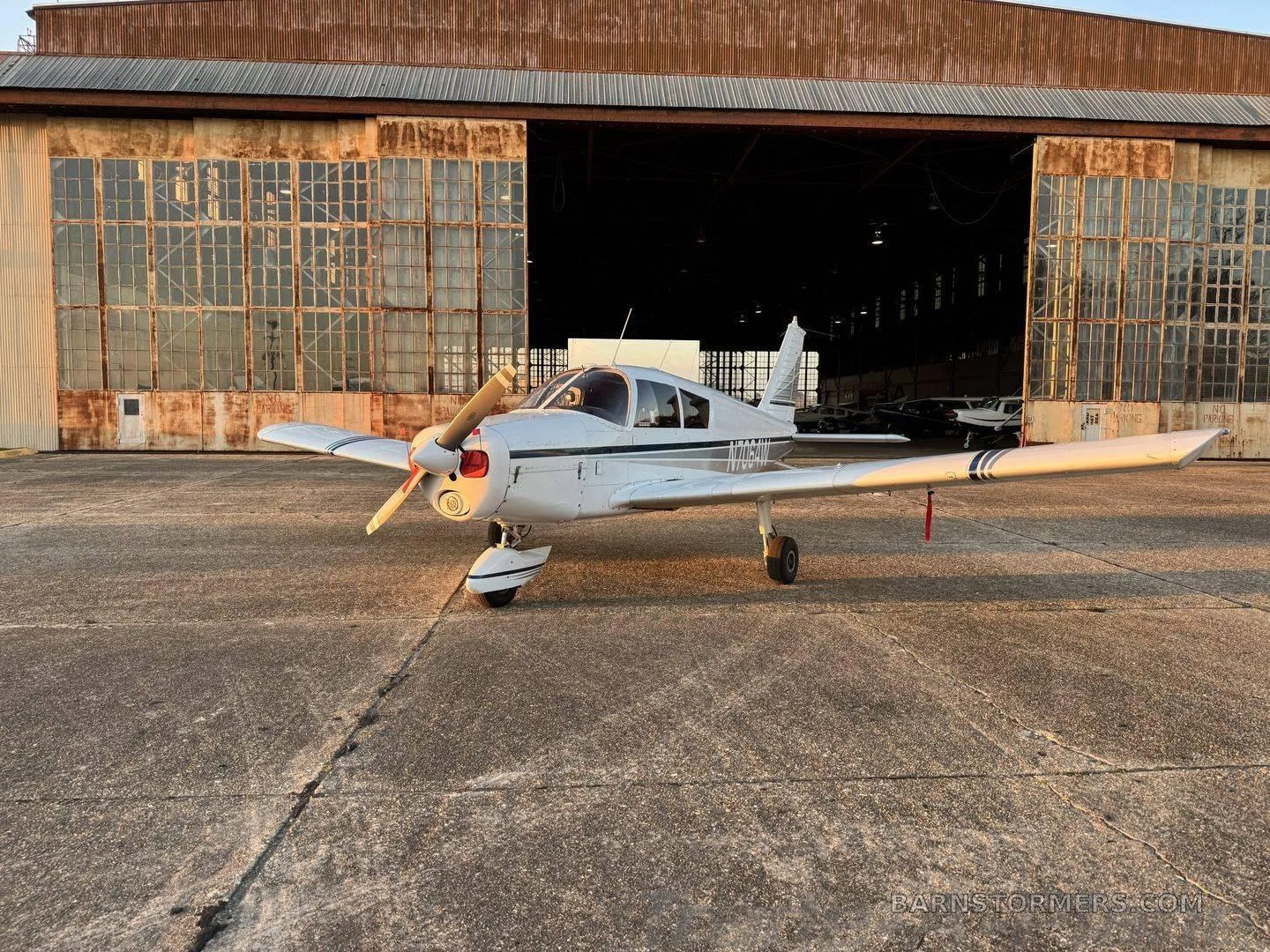 A white single-engine propeller aircraft with dark stripes parked on a concrete ramp in front of an old hangar at sunset.