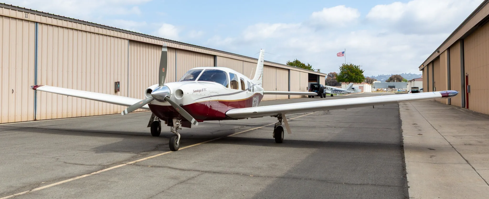 A white and maroon Piper Saratoga II TC aircraft, registration N691MA, parked on an asphalt ramp with hangars and an American flag in the background.