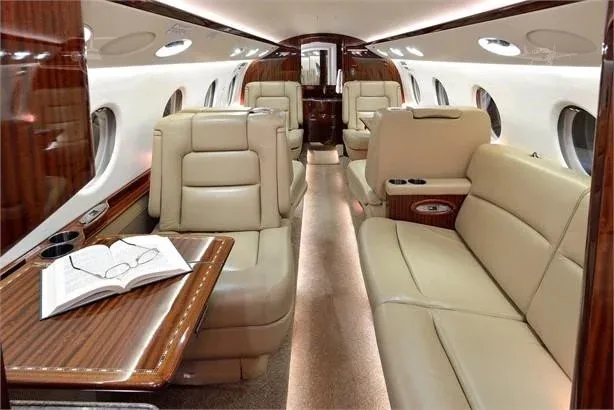 A view of the private jet's cabin, showing beige leather seating, a wooden table with a book, and large windows.