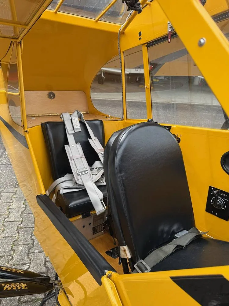 Interior view of a yellow aircraft cabin, showing two black leather seats with grey harnesses and a wooden floor.