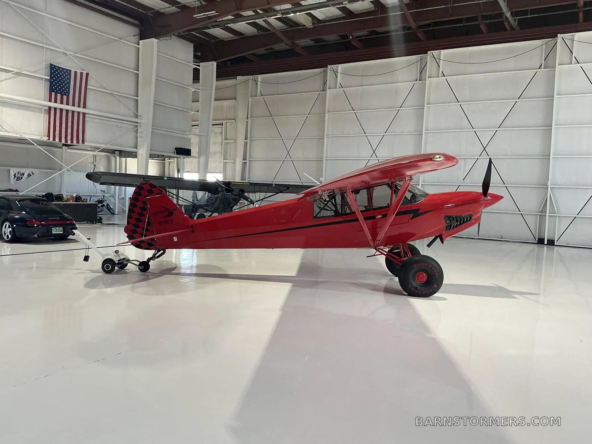 A red Bushmaster aircraft with a checkered tail is parked in a hangar, viewed from the front-left.