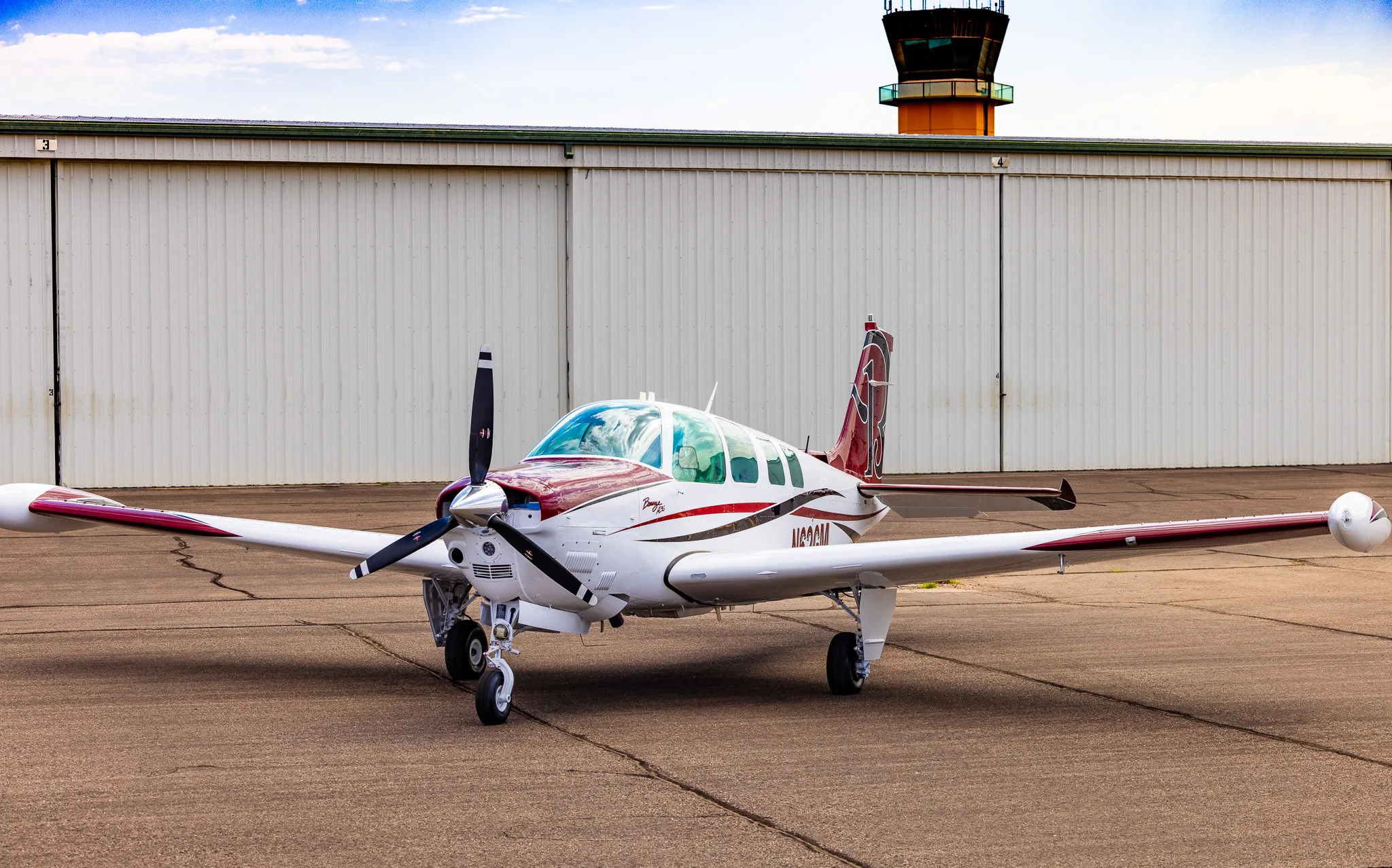 A white and maroon Beechcraft Bonanza A36 is parked on an airport tarmac in front of a hangar with an air traffic control tower in the background.