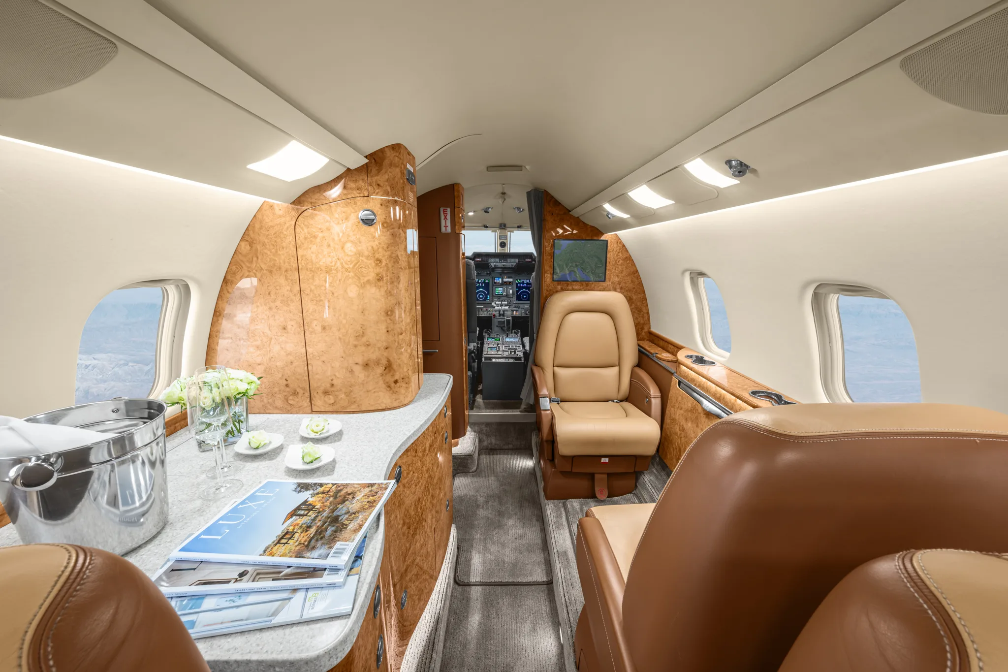 The elegant interior of a private jet cabin with tan leather seats, wood paneling, and a counter with magazines and a champagne bucket, looking towards the cockpit.