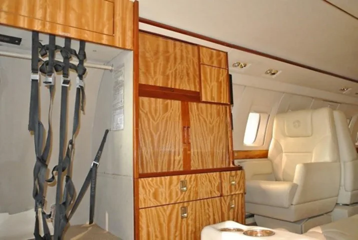 A view inside a private jet cabin showing light wood cabinetry, a single cream leather seat, and safety harnesses.