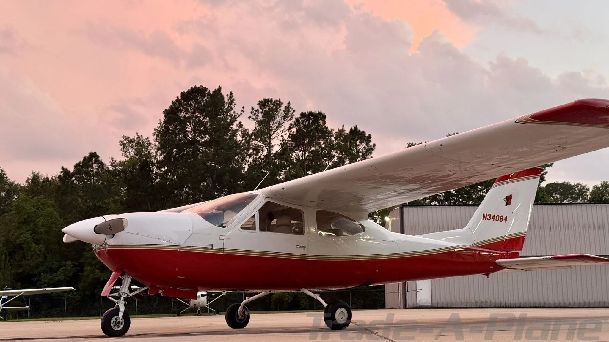 A red and white Cessna 177RG Cardinal with gold stripes parked on an airfield at sunset.