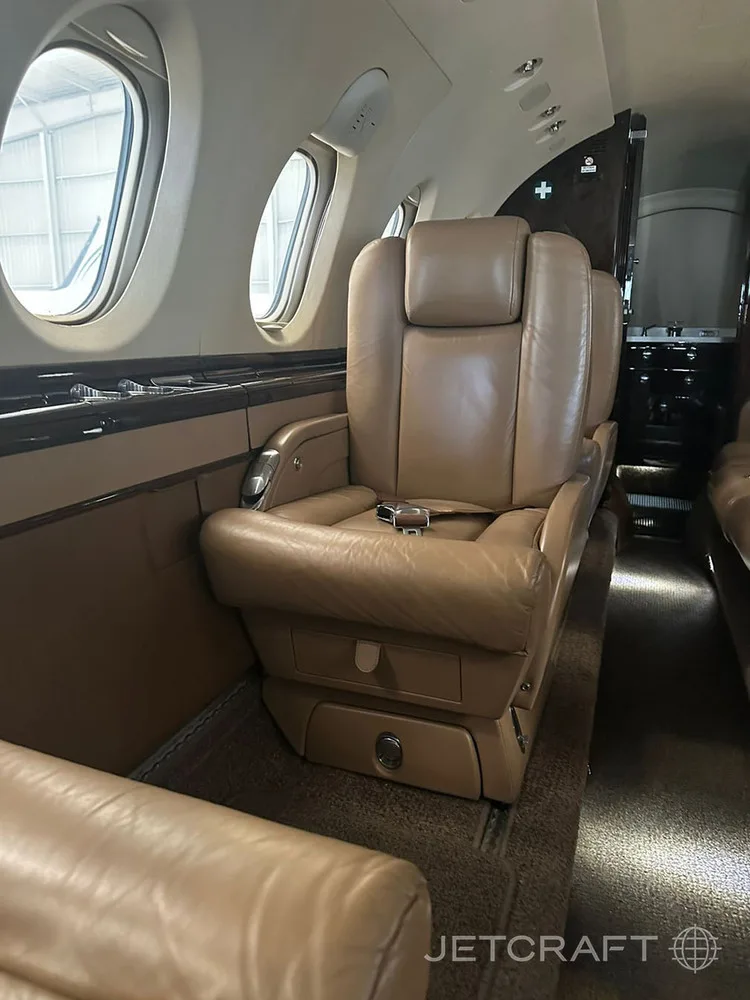 A tan leather single seat in a private jet cabin, with a window and part of the galley visible.