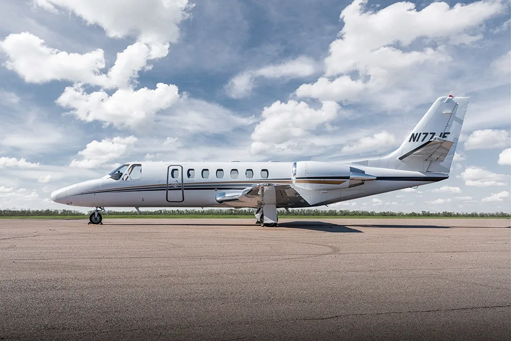 A white Cessna Citation business jet with gold and black stripes is parked on an asphalt tarmac under a partly cloudy sky.