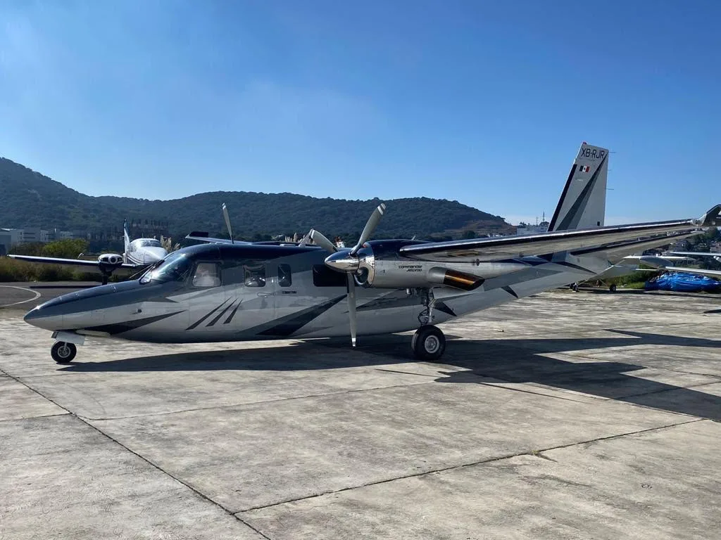 A silver and dark grey Commander 690B twin-engine turboprop, registration XB-RUR, parked on an airfield with mountains in the background.