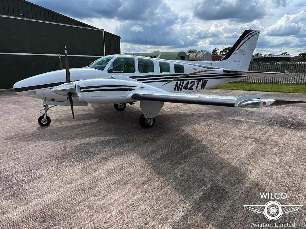 A white Beechcraft Baron N142TW with black and gold stripes parked on a tarmac next to a hangar.