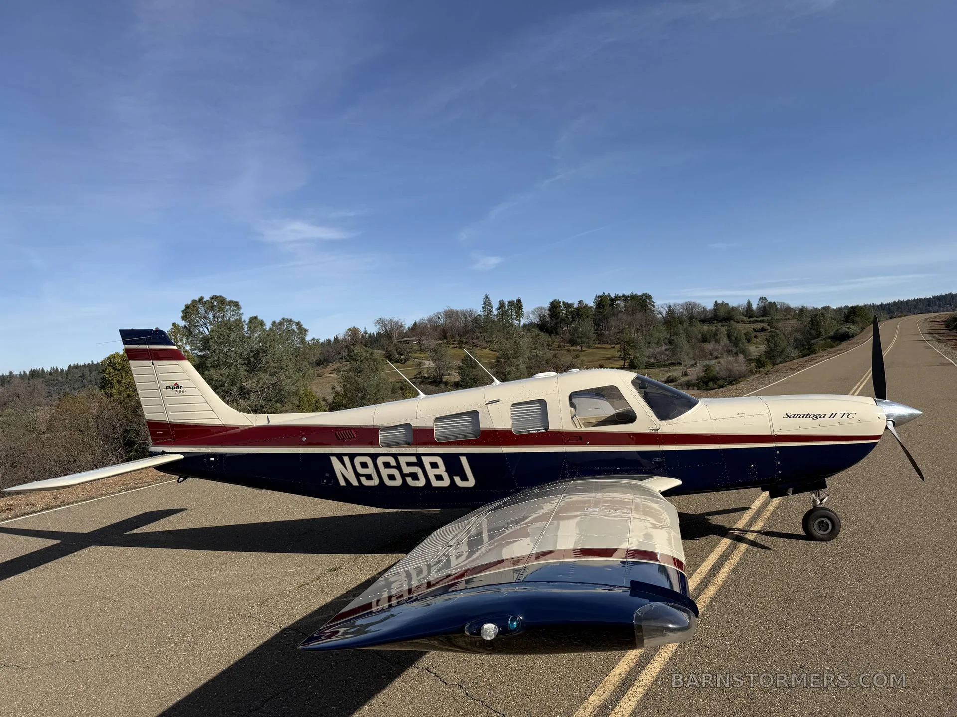 A three-quarter front view of a white, red, and blue Piper Saratoga II TC aircraft (N965BJ) parked on an asphalt surface with trees in the background.
