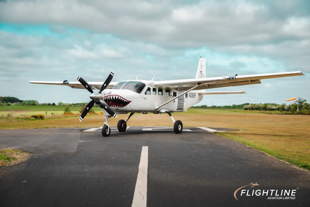 A white Cessna 208 Caravan with shark teeth nose art, parked on a runway under a cloudy sky.