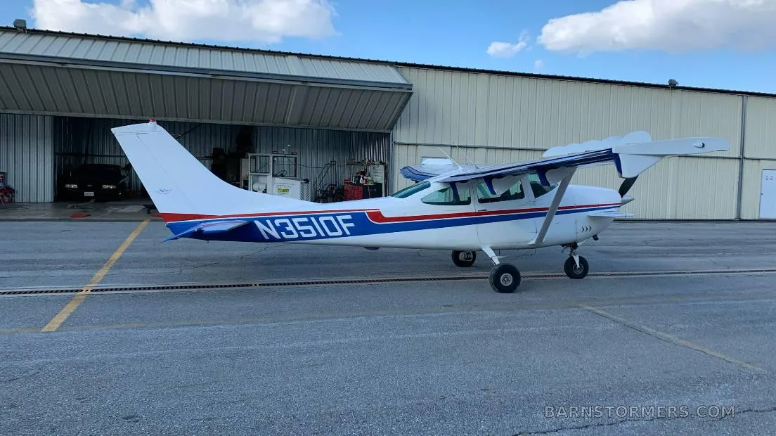 A white Cessna 172 with blue and red stripes, registration N351OF, parked on an asphalt ramp in front of a hangar.