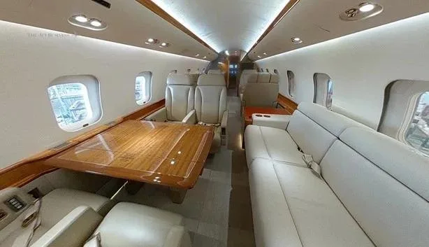 A private jet cabin interior with a beige leather sofa on the right, individual seats, and a large wooden table on the left.