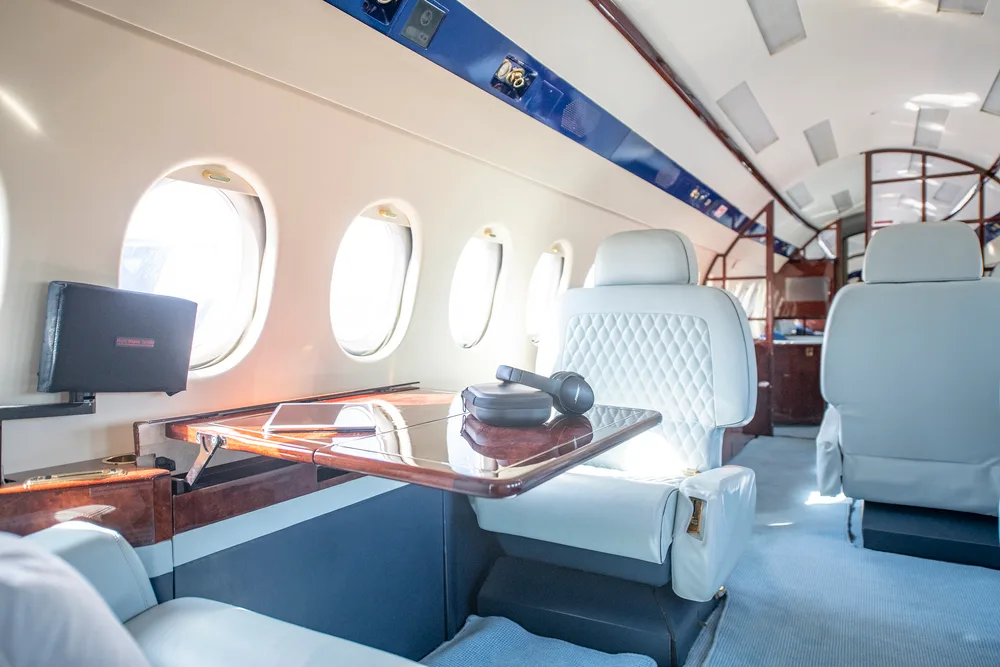 A close-up view of the private jet's cabin interior, showing light grey quilted seats, a wooden table with a tablet and headphones, and large windows.