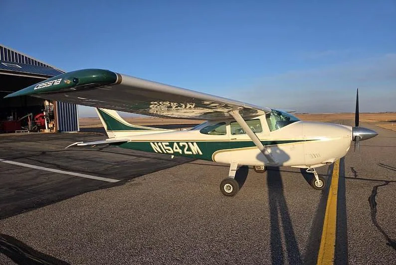 A white and green Cessna aircraft (N1542M) is parked on an asphalt runway next to a hangar under a clear blue sky.