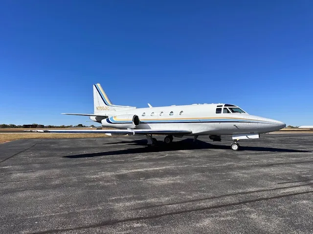A white business jet with blue and gold stripes, registration N700JC, parked on an asphalt tarmac under a clear blue sky.