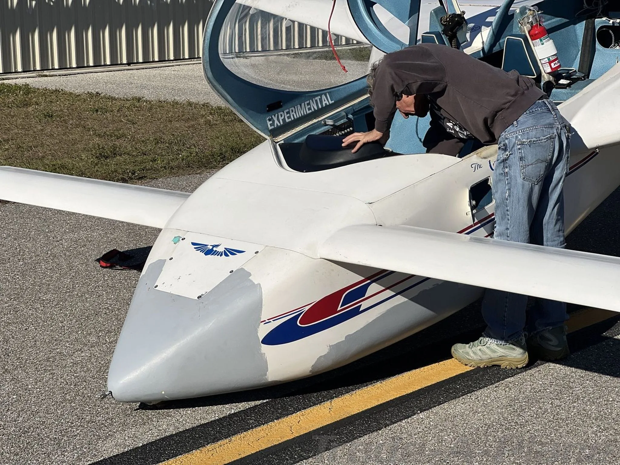 A person leans into the open canopy of a white experimental aircraft with blue and red stripes, parked on an asphalt runway.