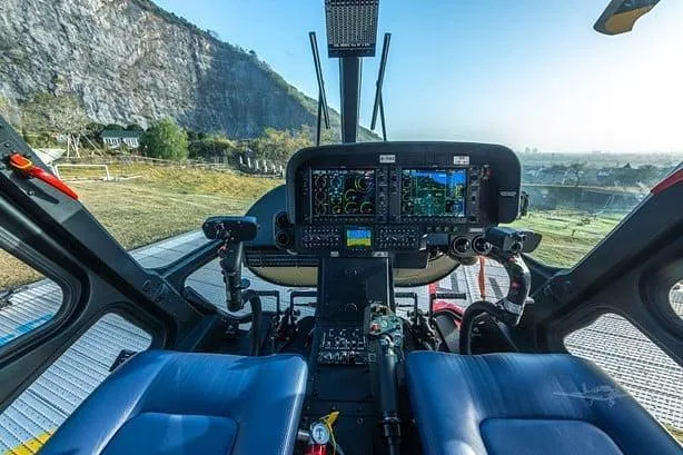 The cockpit of a helicopter features multiple digital displays and controls, with a view of a rocky mountain outside.